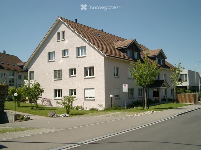 Two-story residential house with beige siding, multiple windows, and a tiled roof. The house is surrounded by a small garden with trees and shrubs.