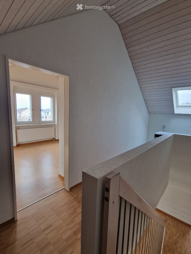 Empty attic room with wooden flooring, slanted wooden ceiling, and a staircase leading to another room. The adjacent room has large windows and a radiator.