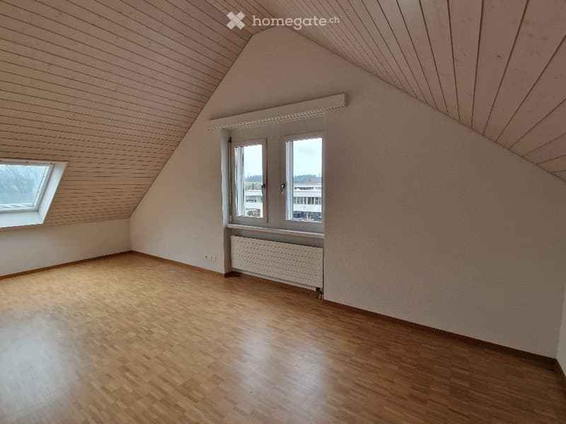 Empty attic room with wooden flooring, white walls, and large windows, including a skylight, offering natural light.