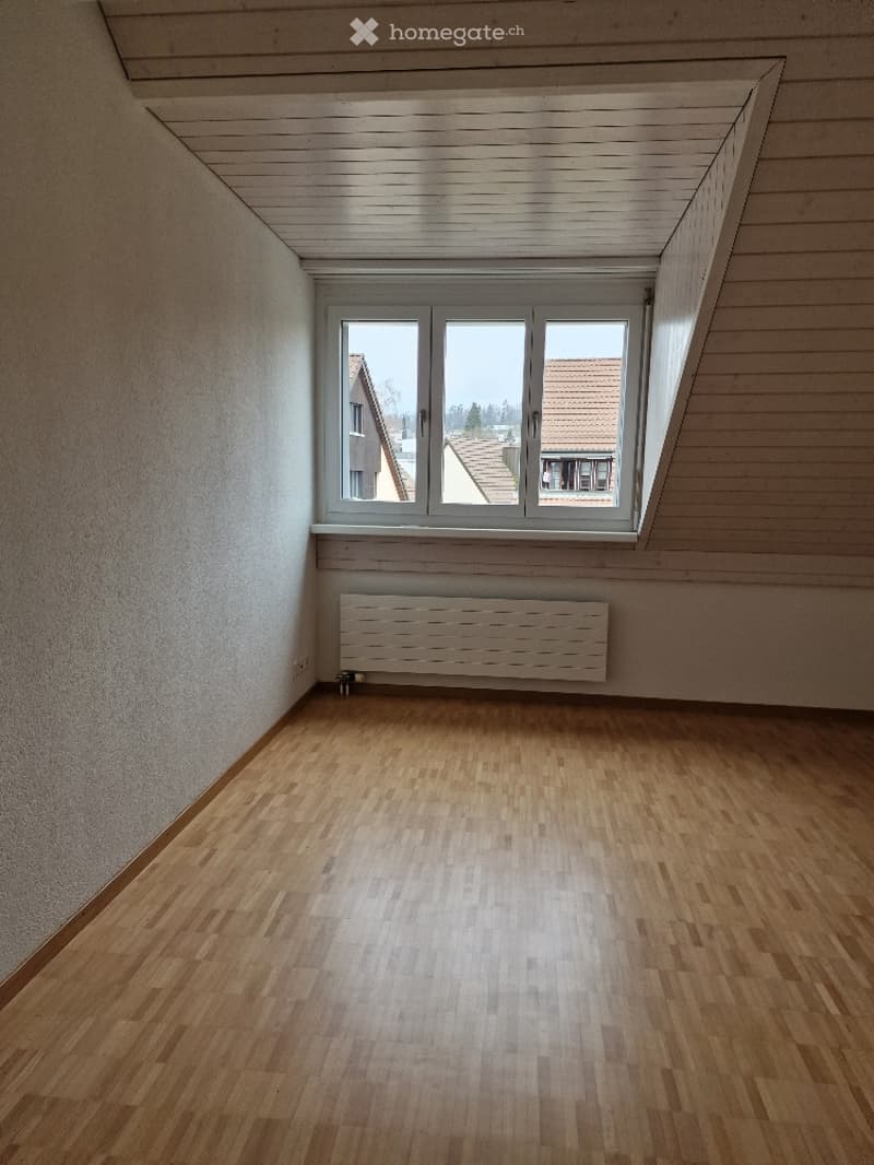 Empty attic room with wooden flooring, white walls, and a large window offering a view of neighboring houses.
