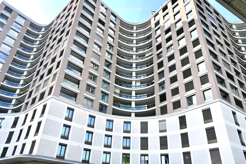 Modern high-rise building with curved balconies and large windows, featuring a mix of grey and white exterior walls and black shutters.