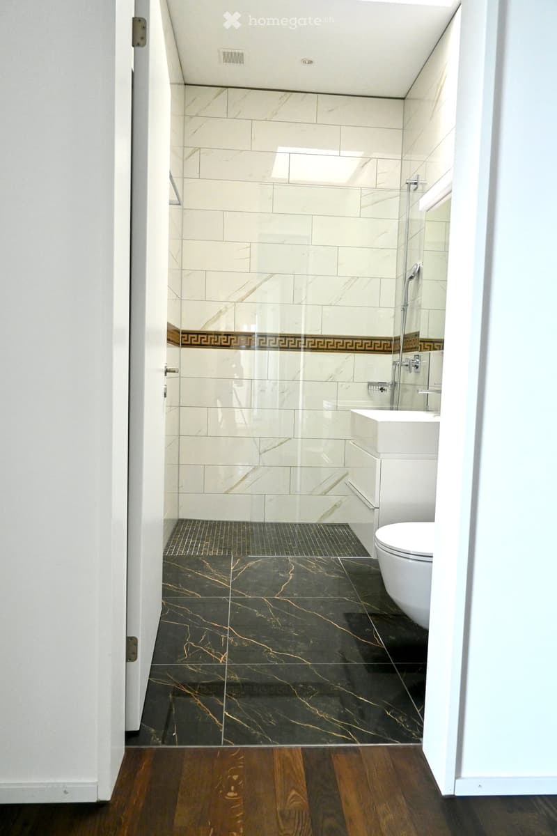 Modern bathroom with white tiles, dark marble flooring, glass shower enclosure, and a white toilet and sink.