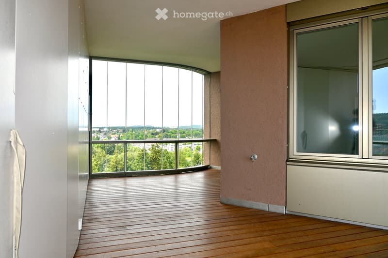 Empty balcony with wooden flooring, large glass windows, and a view of trees and buildings.
