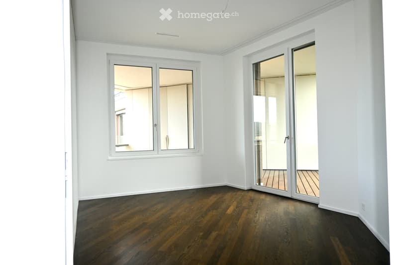 Empty room with white walls, wooden flooring, and large windows. A sliding glass door leads to another room with wooden flooring.