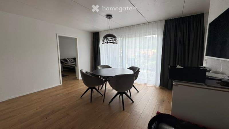 Bright dining area with a round table and four chairs, large windows with sheer curtains, and a modern hanging lamp.