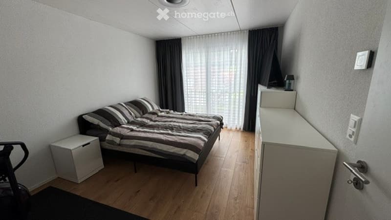 Cozy bedroom with a double bed, striped bedding, a nightstand, and a wardrobe. The room features wooden flooring and large windows with dark curtains.