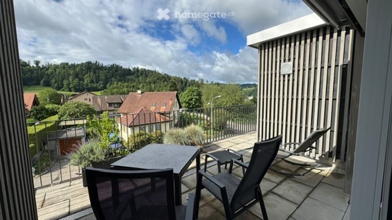 Balcony with outdoor furniture, overlooking a scenic countryside view with houses and greenery.