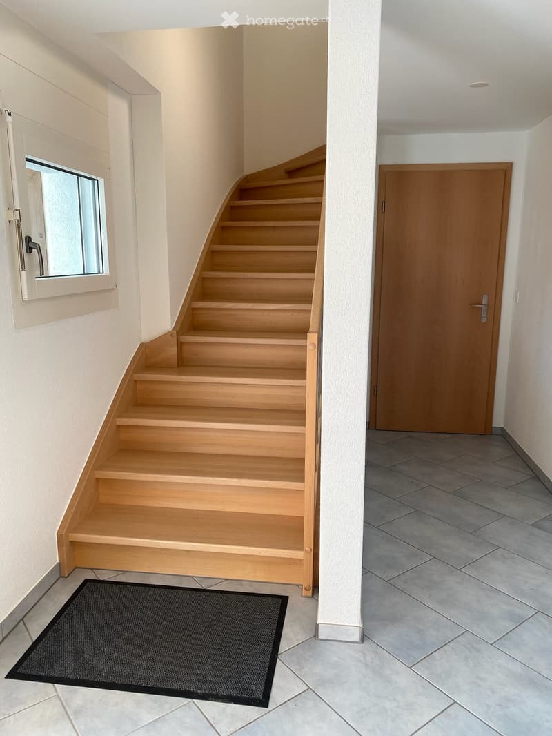 A modern hallway with a wooden staircase, tiled floor, and a black doormat at the base of the stairs. A wooden door is visible on the right side, and a window with a white frame is on the left.