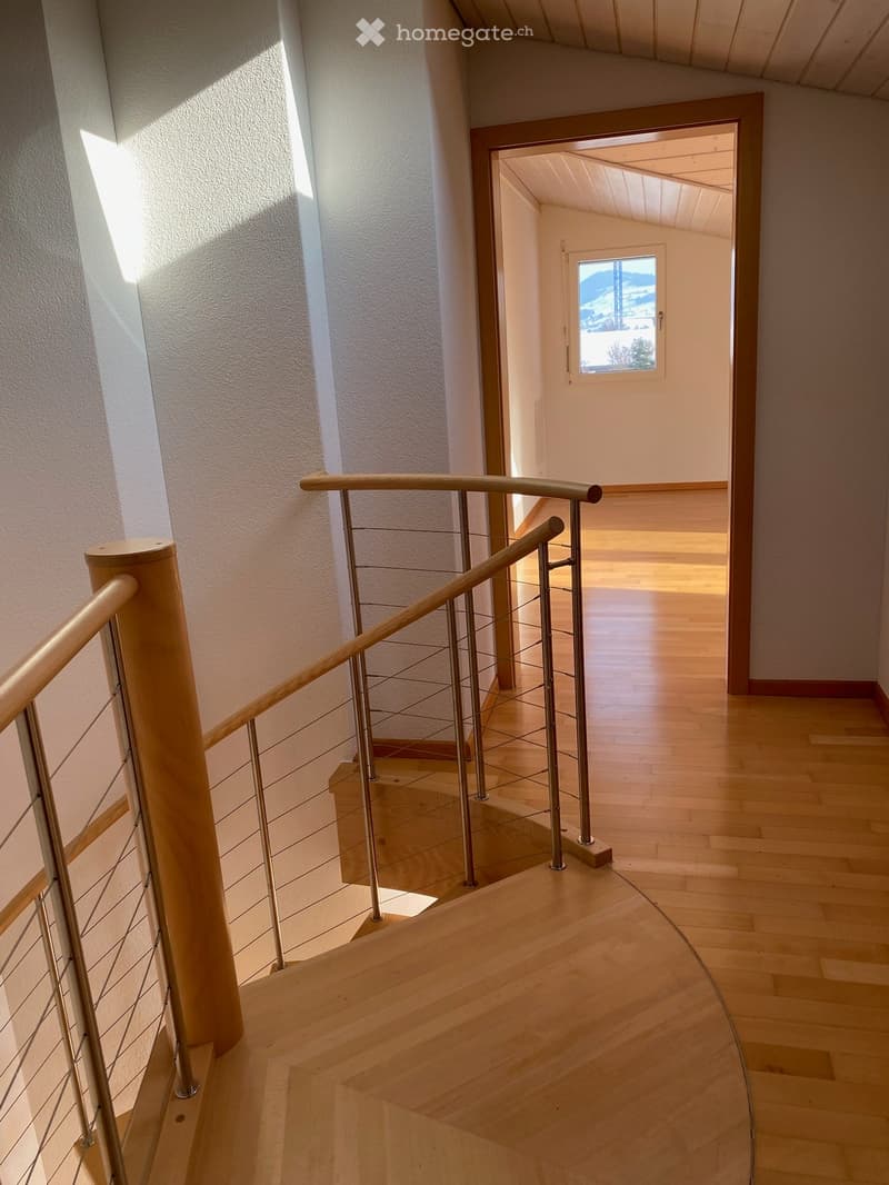 Spacious hallway with wooden flooring, modern staircase featuring a wooden handrail and metal balustrade, leading to a room with a window offering a scenic view.