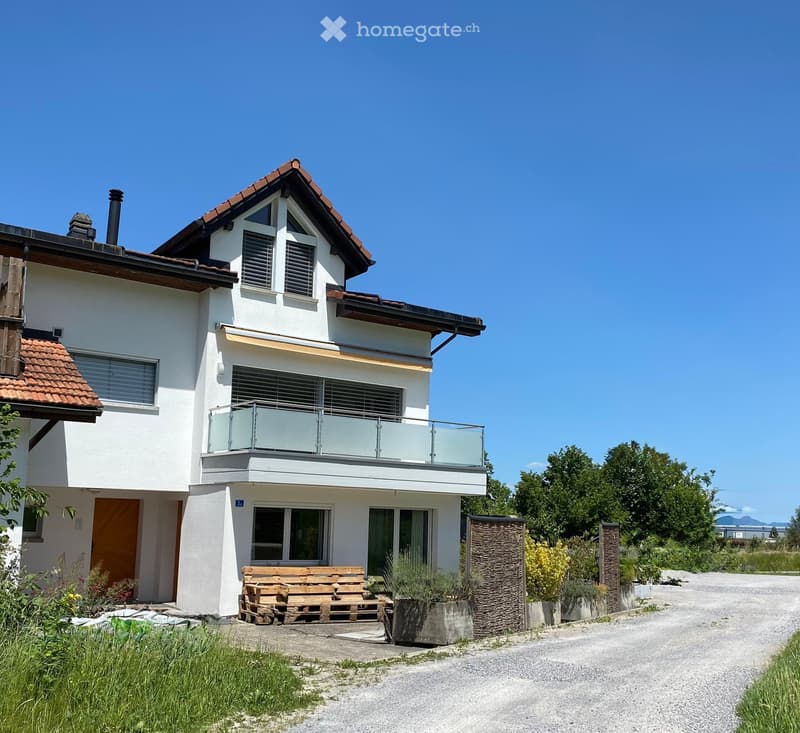 Two-story modern house with white walls, red-tiled roof, large windows, and a balcony with glass railing. The house features a wooden bench and small garden area in front.