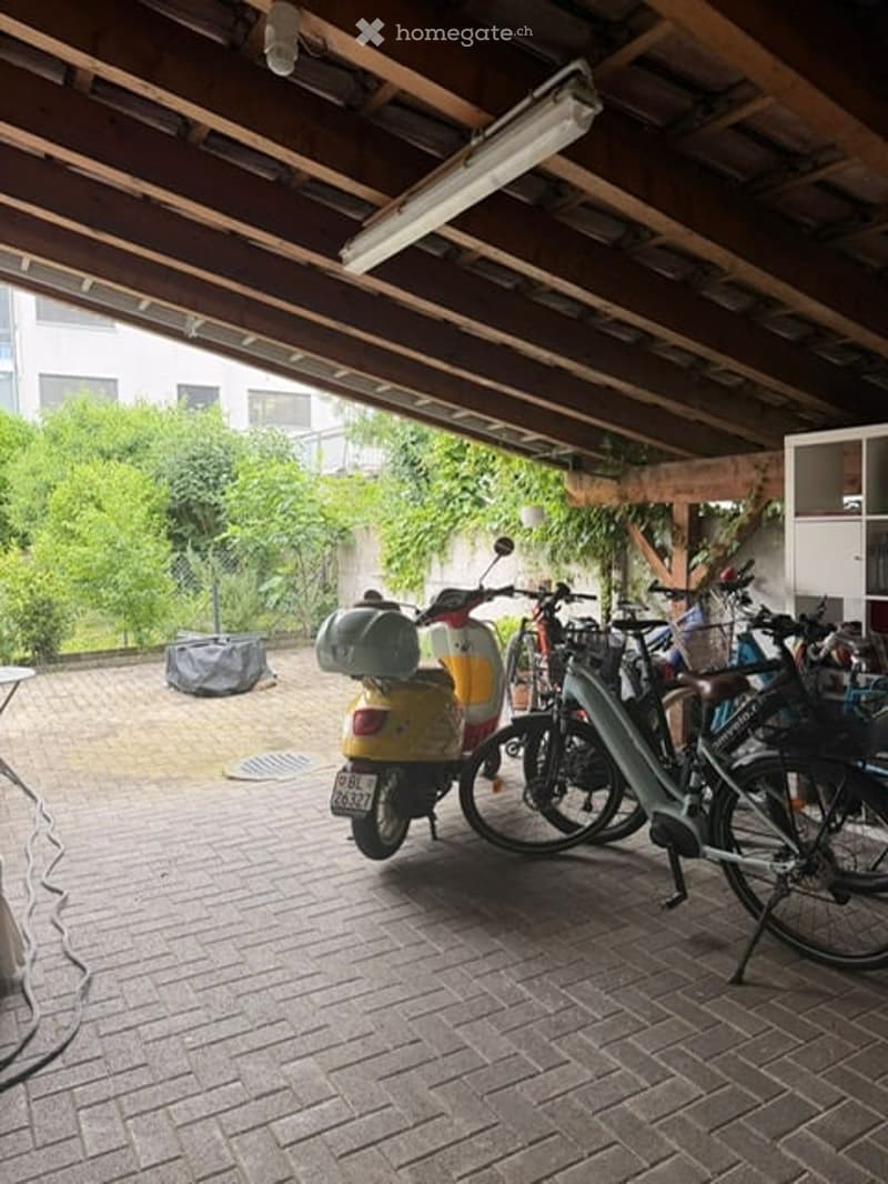 Covered outdoor area with brick flooring, several bicycles and a scooter parked, wooden ceiling beams, and greenery visible in the background.