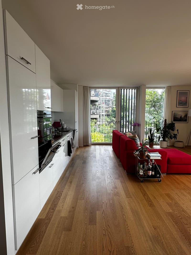 Open-plan living and kitchen area with wooden flooring, white kitchen cabinets, red sofa, small cart with bottles, and large windows leading to a balcony.