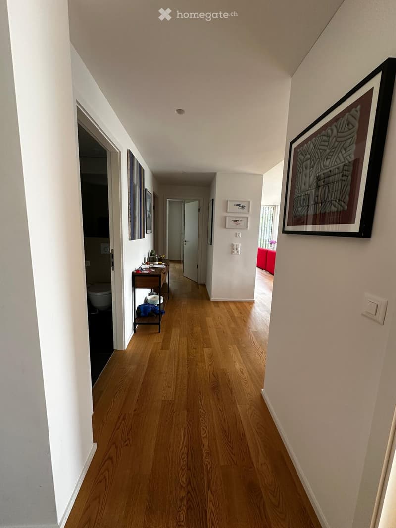 A long, narrow hallway with light wooden flooring and white walls. The hallway features several doors leading to different rooms, framed artwork on the walls, and a small table with items on it to the left. The ceiling has recessed lighting.