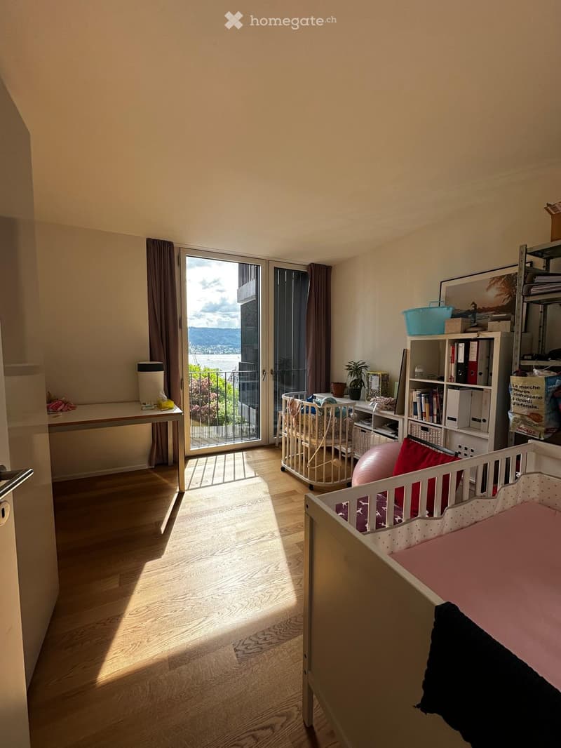 Bright room with wooden floor, large window and balcony, baby crib, desk, shelf, and plant.