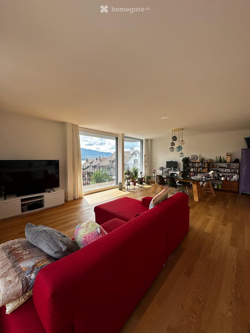 Modern living room with a red sectional sofa, coffee table, TV, dining area with chairs and table, bookshelf, and large windows offering a scenic view.