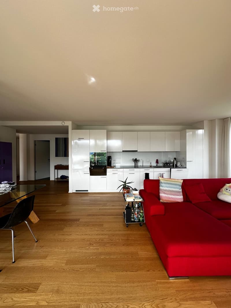 Modern living room with a red sectional sofa, coffee table, glass dining table, and a small kitchen area with white cabinets and stainless steel appliances.