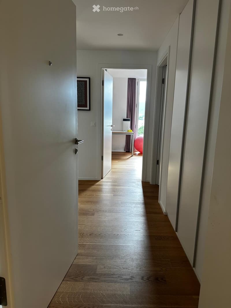 A long, narrow hallway with wooden flooring and white walls, leading to an open doorway with a small table and a red exercise ball visible in the adjacent room.
