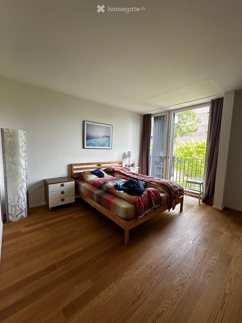 Bedroom with wooden flooring, a bed with a colorful quilt, a nightstand with a lamp, a picture frame on the wall, and a small stool near the balcony.