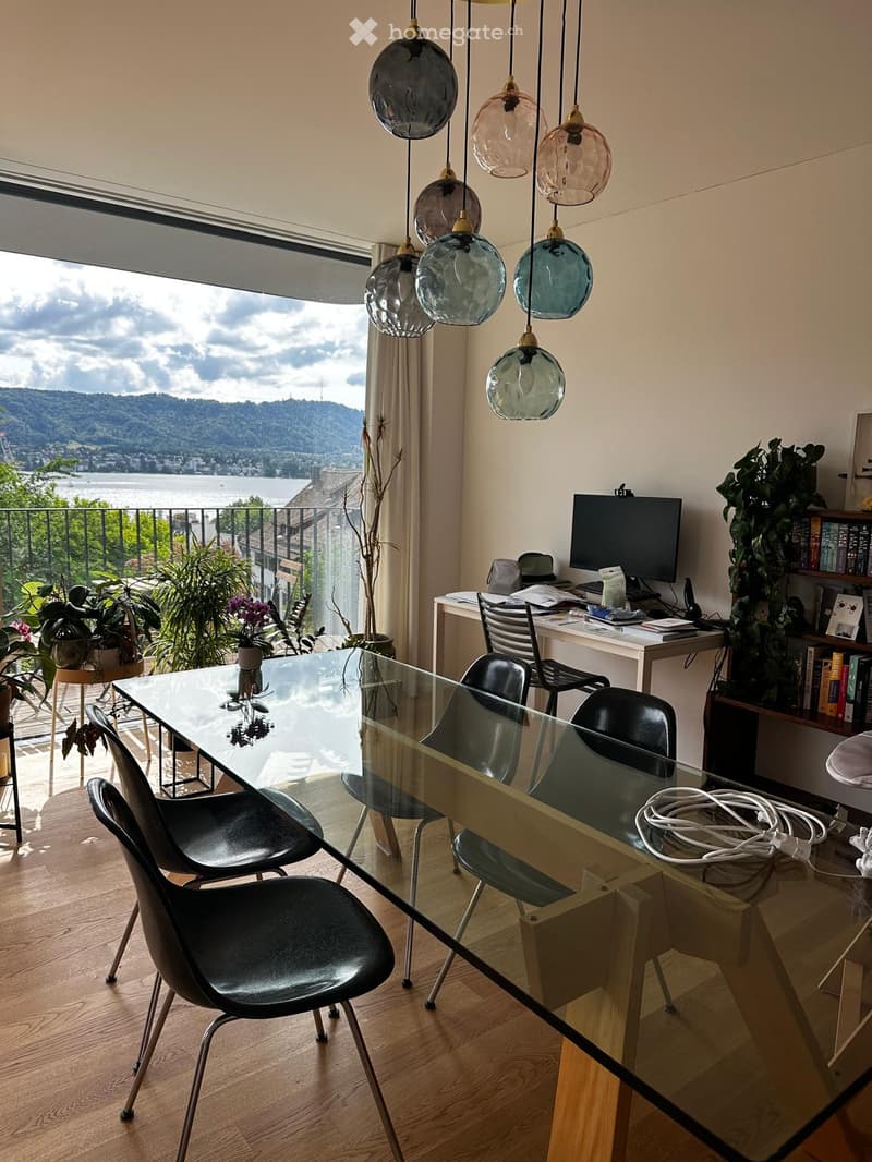 Modern dining area with a glass table, black chairs, and a view of a lake and mountains through large windows. There is a desk with a laptop and a bookshelf with books and plants.