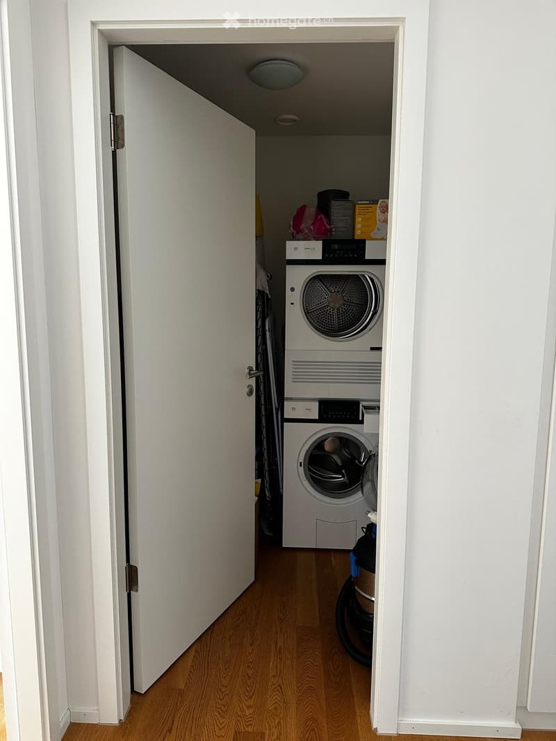 A narrow hallway leads to a small laundry area with a washing machine and dryer, wooden flooring, and white walls.