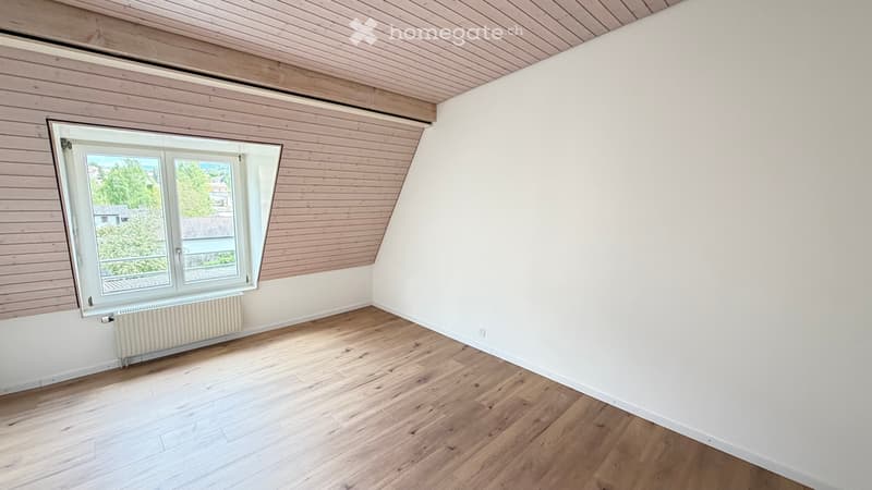 Empty room with wooden flooring, white walls, and a large window offering a view of greenery outside. The ceiling has wooden paneling, and there is a radiator below the window.