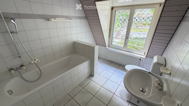Bright bathroom with white tiled walls and floor, featuring a bathtub, sink, and toilet. A large window offers a view of the outdoors.
