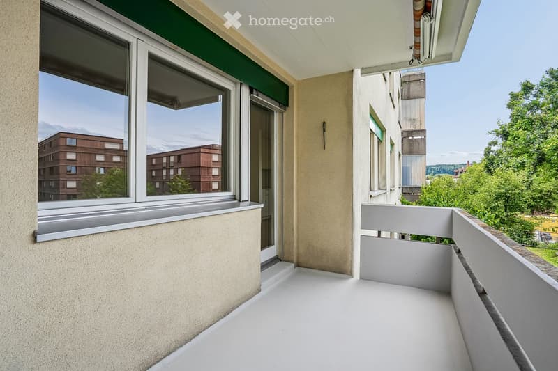 Balcony with white floor, large windows, and a view of surrounding buildings and greenery.