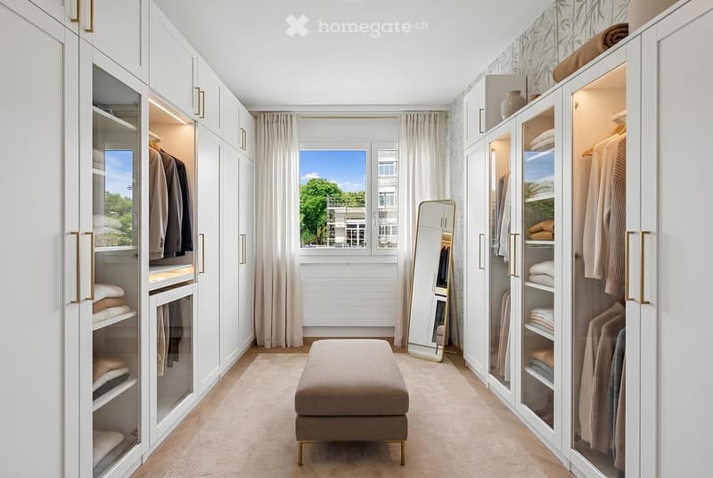 Well-organized walk-in closet with white built-in wardrobes, neatly arranged clothing, folded towels, and a small beige stool in the center.