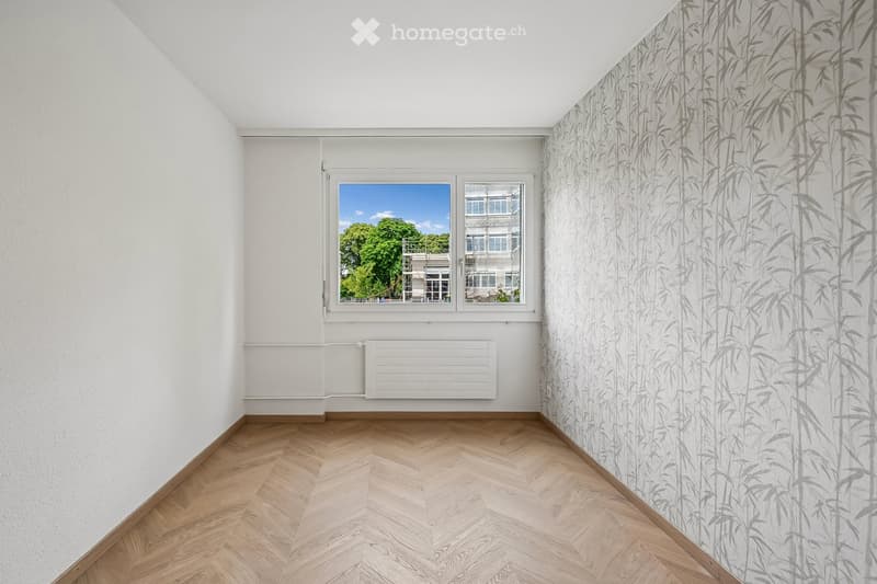Empty room with light wooden herringbone flooring, white walls, large window with view of greenery, and a radiator below the window.