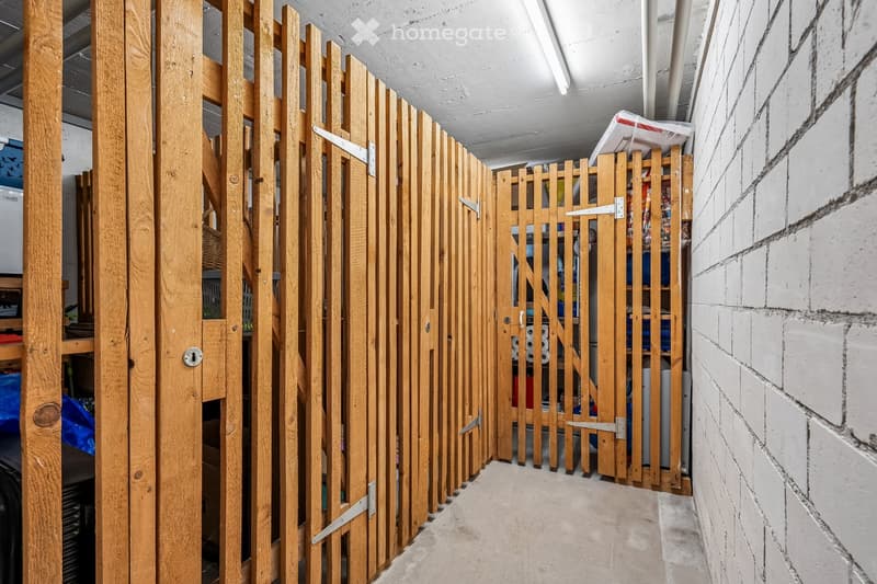A narrow hallway with wooden doors and shelves, concrete floor, white brick wall, and storage items visible behind the doors.