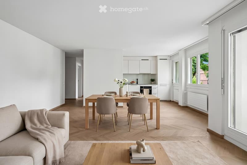 Bright, open-plan living and dining area with light wood flooring, white walls, and large windows. Features a sofa with a throw, coffee table with decorative items, dining table with chairs, and a modern kitchen area with white cabinets and stainless stee