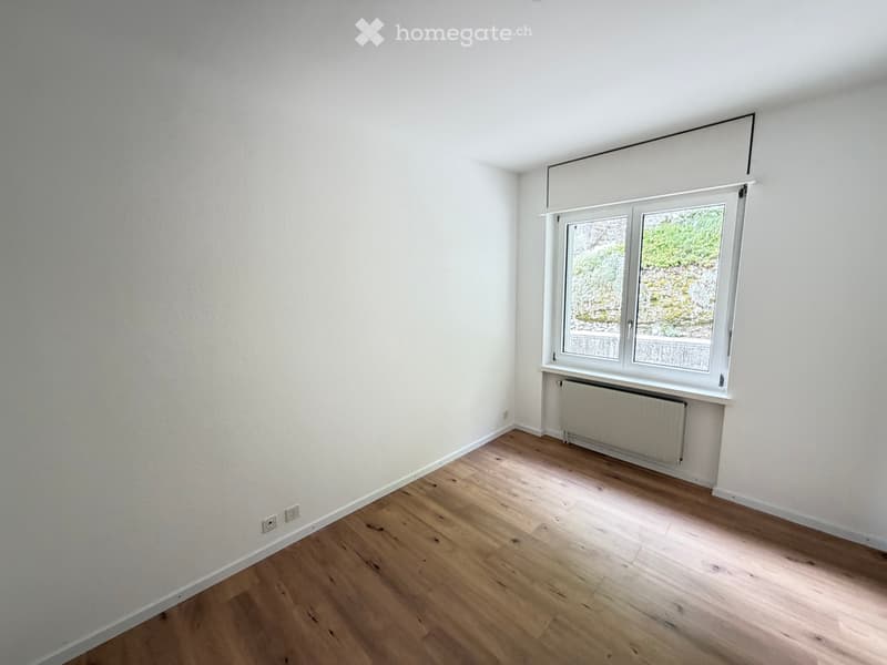 Empty room with light wooden flooring, white walls, and a large window with a radiator below it.