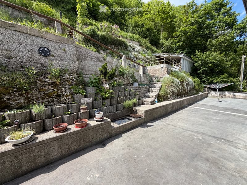 Garden area with stone steps, potted plants, and a small water feature, surrounded by greenery and a stone wall.