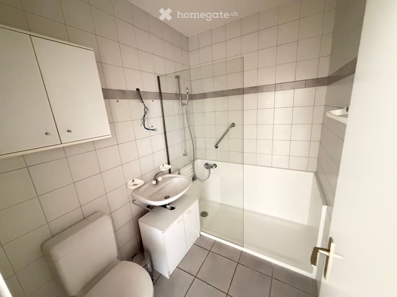 Clean, modern bathroom with white tiles, a toilet, a sink with a cabinet, and a walk-in shower with a glass door and grab bar.