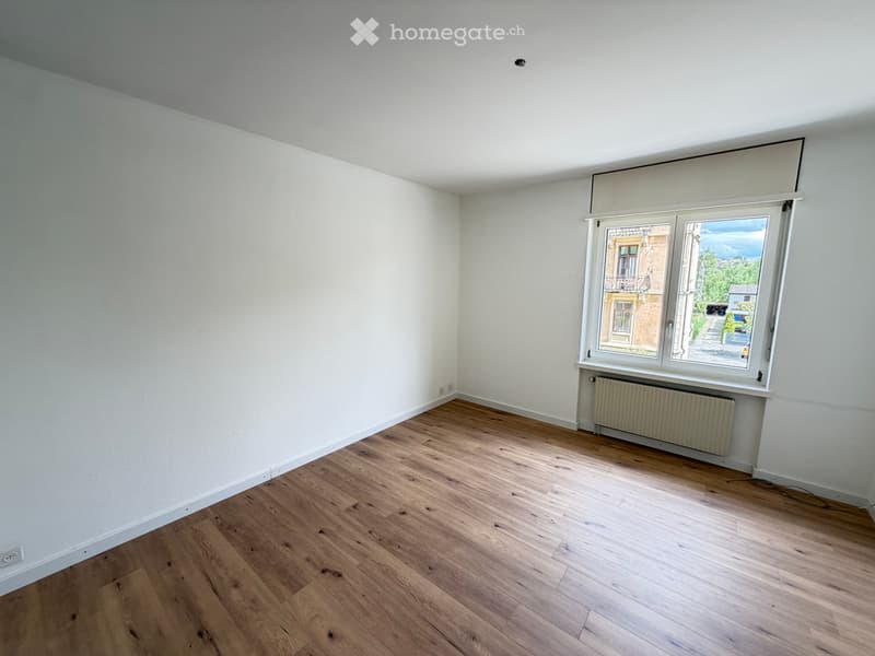 Empty room with wooden flooring and white walls, featuring a large window with a view of the street and a radiator below it.