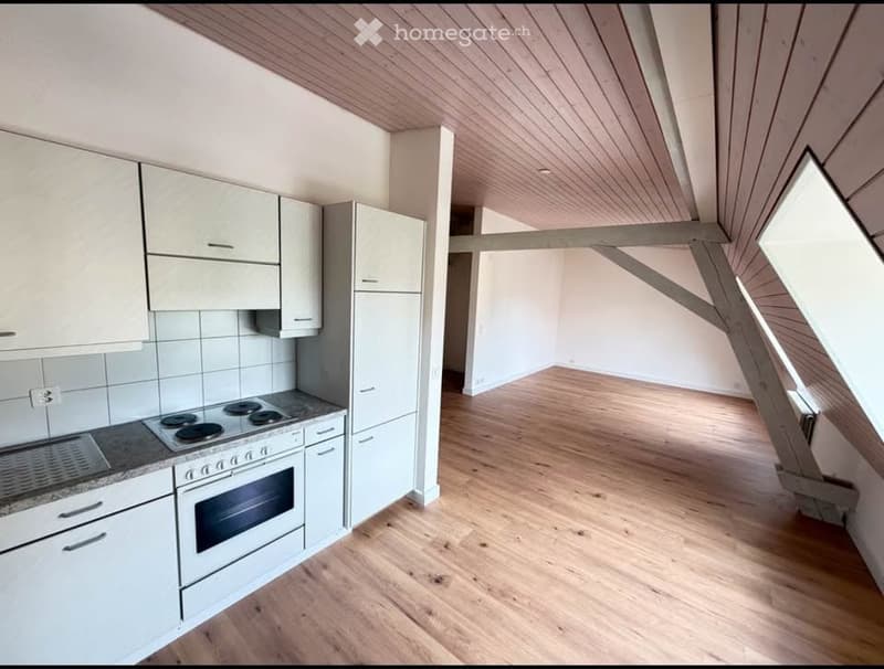 Bright, modern kitchen with white cabinets, a stove, and an oven, connected to a spacious living area with wooden flooring and exposed wooden beams on the ceiling.