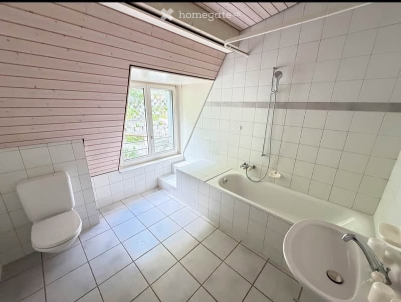 Bright bathroom with white tiled walls and floor, featuring a toilet, bathtub, sink, and a window offering a view.