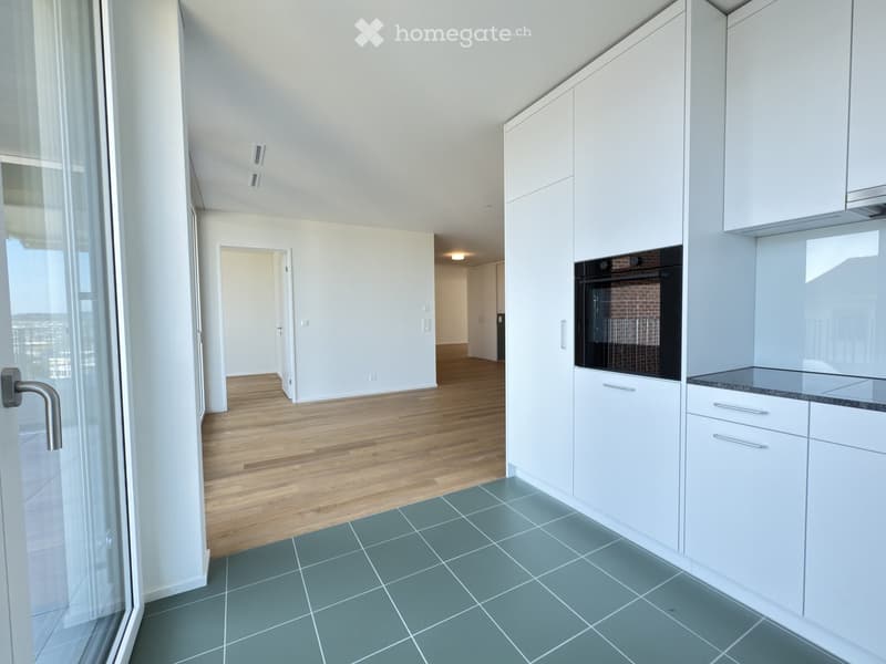 Modern unfurnished kitchen with white cabinets, stainless steel oven, and grey tiled floor. Open-plan layout with wooden flooring in adjacent area and large windows offering a view.