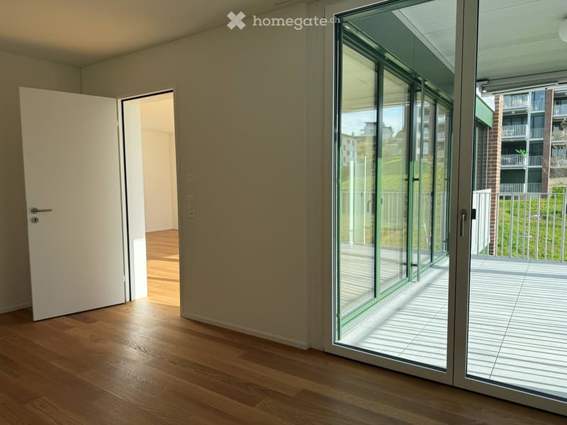 Empty room with wooden flooring, large glass sliding doors leading to a balcony, and an open doorway to another room.