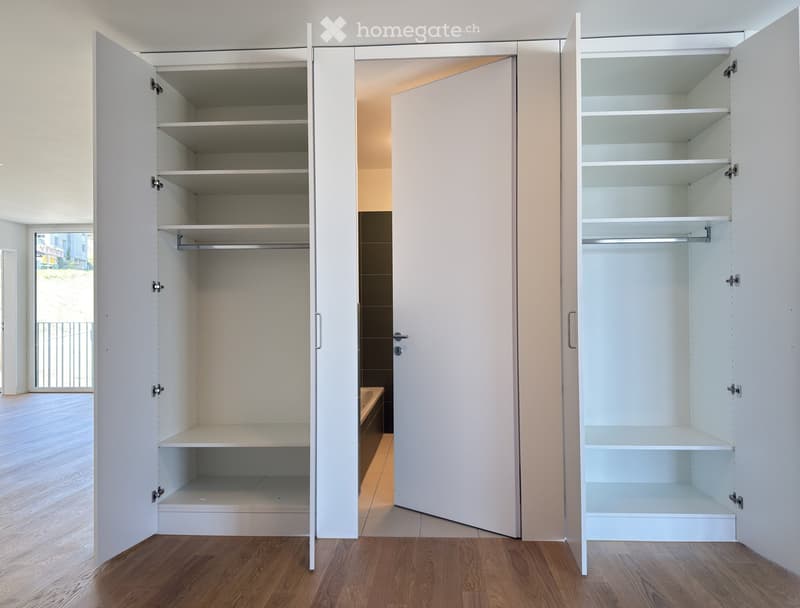 Empty room with white built-in wardrobes and shelves, wooden flooring, and a view of a bathroom area through an open doorway.