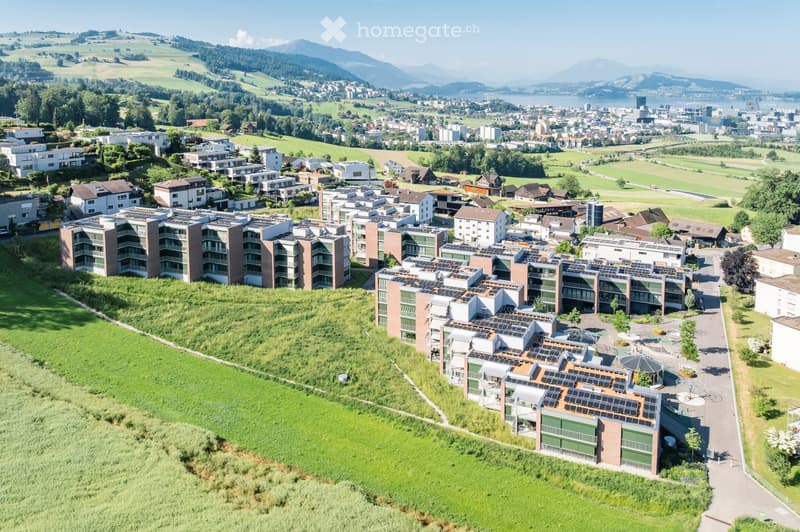 Aerial view of a modern residential complex with multiple apartment buildings, solar panels on rooftops, green spaces, and surrounding landscape with hills and a lake in the background.