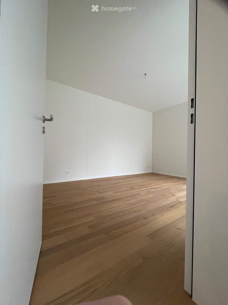Empty room with light wooden flooring and white walls, featuring a door frame and visible electrical outlet.