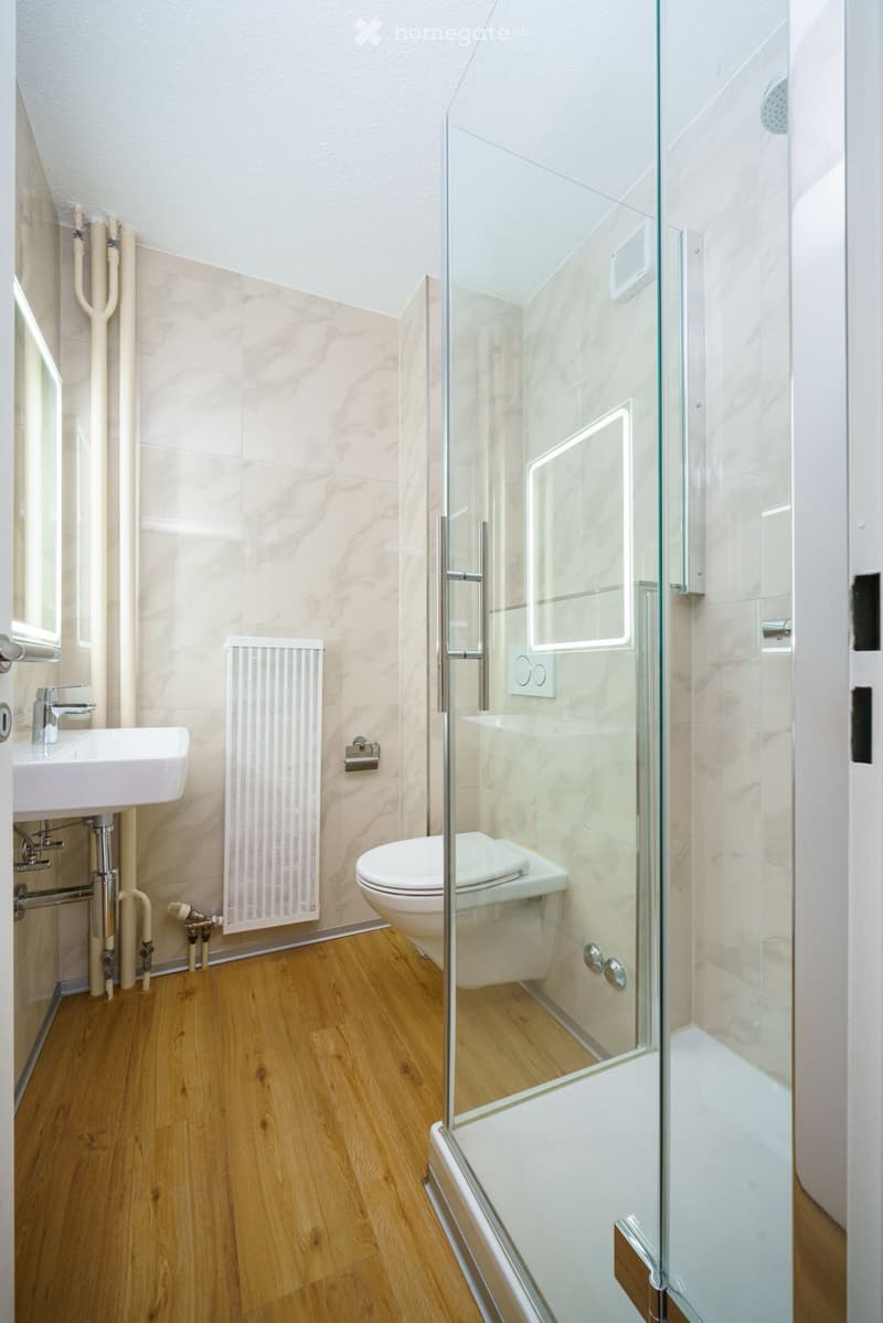 Modern bathroom with light wooden flooring, marble tiles, a white sink, toilet, and a glass-enclosed shower with a modern design.