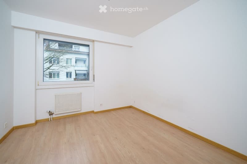 Empty room with light wooden flooring, white walls, a single window showing a neighboring building, and a radiator below the window.
