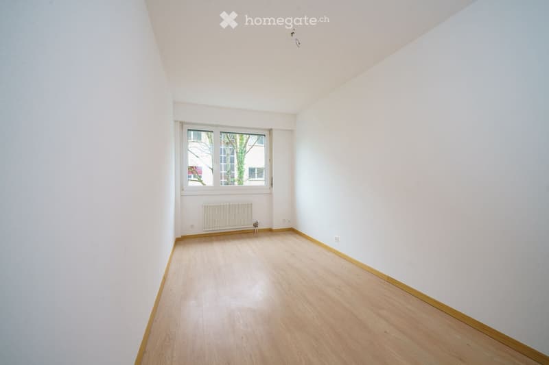 Empty room with light wooden flooring, white walls, and a single window with a radiator below it.