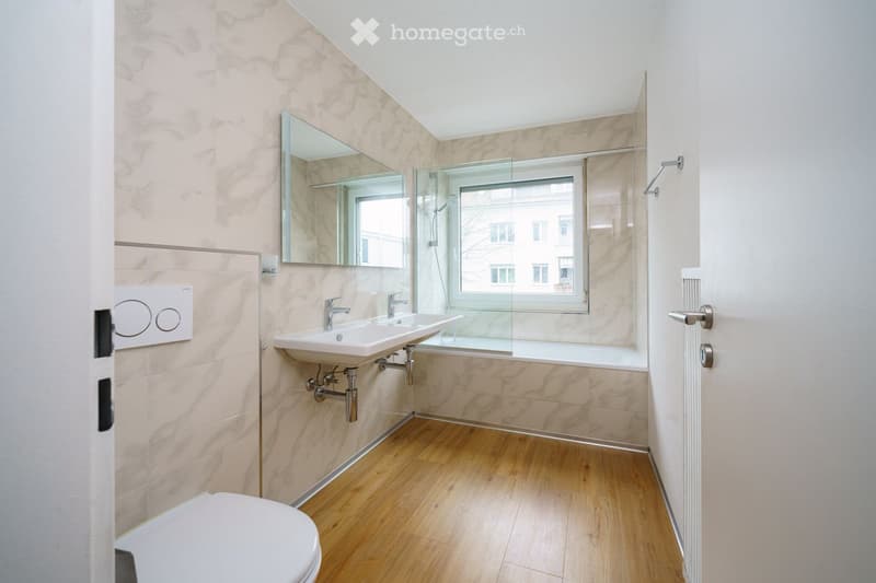 Modern bathroom with marble tiles, wooden floor, double sink, toilet, large mirror, and a window overlooking a cityscape.
