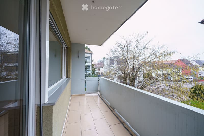 Balcony with tiled floor, white railing, and a view of the surrounding area with trees and buildings.