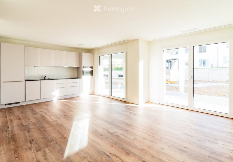 Empty room with light wooden flooring, white walls, and large sliding glass doors leading to an outdoor area. Kitchen cabinets and appliances are visible in the background.