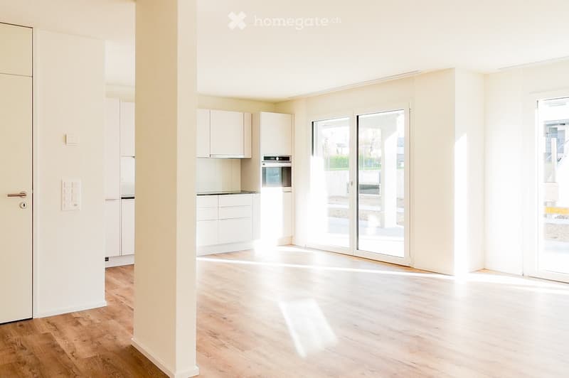 Bright, empty room with wooden flooring, white walls, and large glass sliding doors leading to an outdoor area. The room features a small kitchen area with white cabinets and a built-in oven, but no furniture or appliances are present.