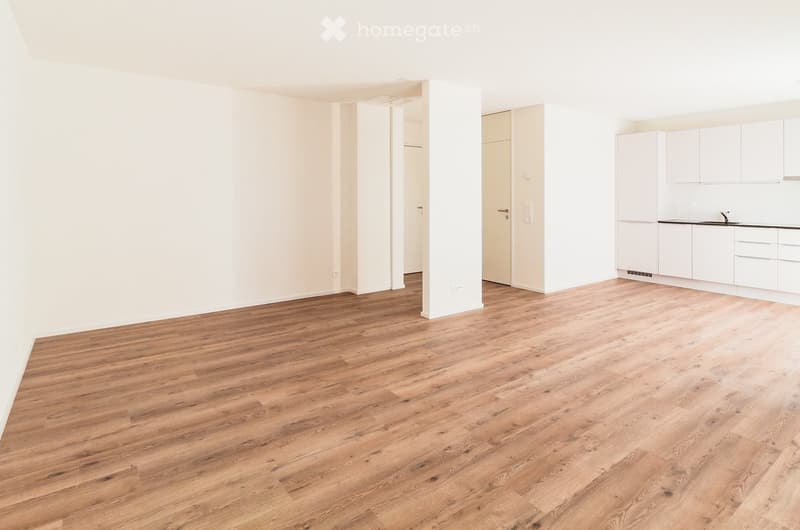 Empty room with light wooden flooring and white walls, featuring a small kitchen area with white cabinets and a sink.